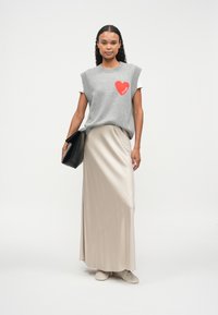 Pull sans manches gris avec un cœur rouge et le texte "FUTURE", associé à une jupe maxi en satin beige et des baskets blanches, tenant un sac à main noir.
