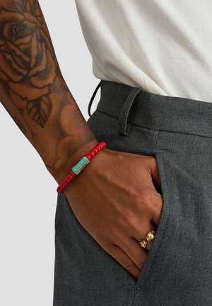 RØdt perlearmbånd med en turkis rektangulær charm indgraveret med "AMOUR", båret på en hånd mod grå bukser.