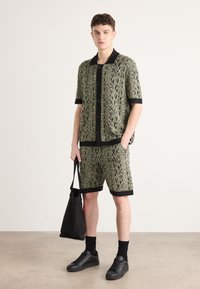 Short-sleeve button-up shirt and shorts in green and black patterned fabric. Black accents at collar, sleeves, and hem. Model holding a black tote bag.