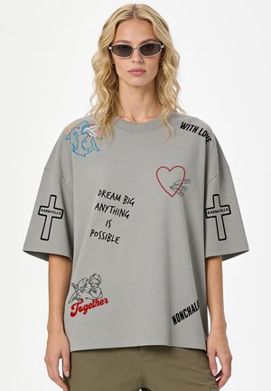 Donna con lunghi capelli biondi indossa una T-shirt grigia oversize con testi e stampe grafiche, inclusi i crocifissi "Dream big anything is possible" e "Karma kills".