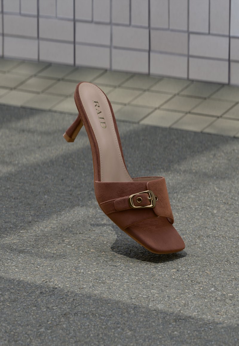 Mule en cuir marron à talon avec boucle dorée et bout carré, debout sur un trottoir gris près d'un mur carrelé.