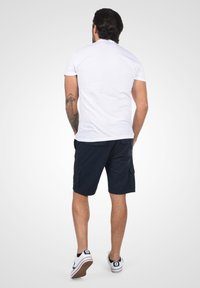 T-shirt en coton blanc, shorts cargo navy, baskets décontractées, tatouage visible sur le bras, vu de dos contre un fond gris clair.