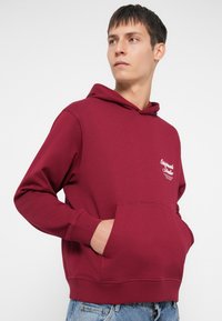 Burgundia kapuutsiga sweatshirt ees oleva taskuga, soonikuotste ja väikese valge logoga vasakul rinnal. Sile kangas.