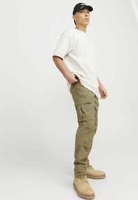 Helles beiges Übergröße-T-Shirt, olivgrüne Cargo-Hosen mit mehreren Taschen und beige Schnürstiefel, vor einem schlichten weißen Hintergrund.