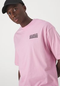 Camiseta de algodón rosa con el texto "Los Angeles Dodgers" en negro en el pecho. Mangas cortas y corte relajado. Llevada con una gorra negra.