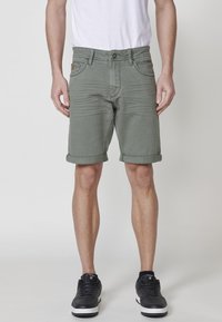 Shorts en denim verts avec des ourlets retroussés, cinq poches et une fermeture éclair. Le tissu a une légère élasticité, offrant un look ajusté. Des baskets noires complètent la tenue.