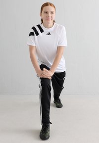 Weißes sportliches T-Shirt mit schwarzen Streifenakzenten und Logo, kombiniert mit schwarzen Hosen mit weißen Seitenstreifen und sportlichen Sneakers.