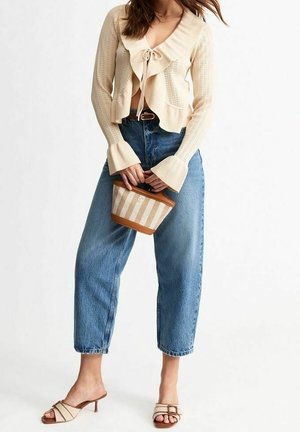 Vrouw draagt een crème gebreid vest, hooggetailleerde blauwe spijkerbroek, beige sandalen met hak, houdt een kleine beige en bruine gestreepte handtas vast.