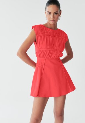 Model poseert zelfverzekerd in een helder rode jurk met een aansluitende lijfje en een uitlopende rok, tegen een effen lichte achtergrond.