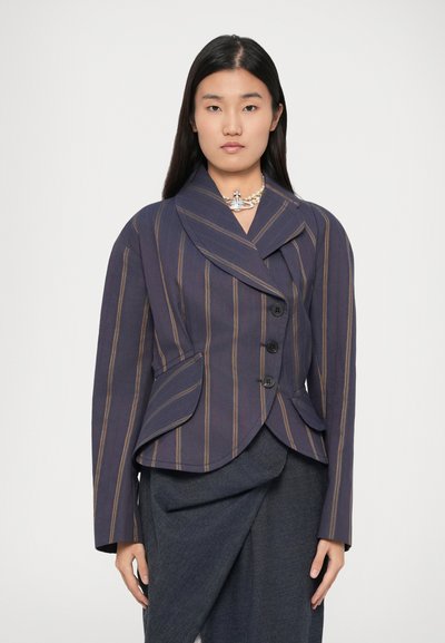 Kvinde med glat sort hår iført en marineblå blazer med brune lodrette striber, en perlekæde med en sølvvedhæng og en mørkegrå nederdel.