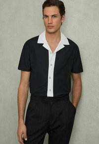 Cămașă neagră cu mânecă scurtă și guler alb, cu dungi verticale, având nasturi pe față și purtată cu pantaloni negri cu dungi.
