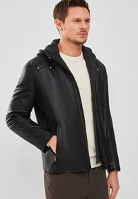 Veste en cuir noir avec une capuche grise, fermeture éclair et deux poches latérales. Texture lisse avec des coutures ton sur ton et des accents matériels minimalistes.