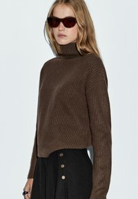 Donna che indossaun maglione a collo alto a coste marrone scuro, una gonna nera con bottoni dorati, occhiali da sole scuri e un anello sulla mano sinistra, su uno sfondo bianco.
