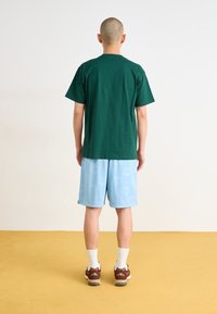 Grünes Baumwoll-T-Shirt, lässiger Schnitt, kombiniert mit hellblauen Shorts mit einem dezenten Muster, braunen Sneakers und weißen Knöchelsocken.
