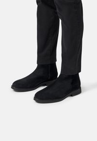 Bottines Chelsea en daim noir avec une finition lisse, fermeture éclair et semelle en caoutchouc noir, portées avec un pantalon noir tapered.