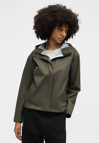 Göndör hajú nő olíva zöld kapucnis dzsekiben és fekete nadrágban, sima világos háttér előtt állva.