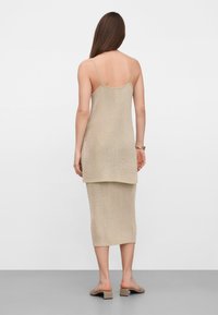 Woman standing facing away wearing a beige sleeveless knit top and matching midi skirt with beige heeled mules on a plain background.