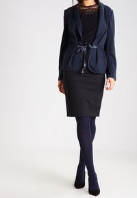 Námořnický blazer s vázáním v pase přes černé šaty, doplněný černými vysokými podpatky a námořnickými punčochami. obsahuje krajkové detaily na blůze.