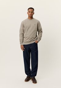 Ανοιχτό γκρι φούτερ με στρογγυλή λαιμόκοψη και κεντημένο λογότυπο, συνδυασμένο με σκούρο μπλε φαρδιά τζιν και καφέ παπούτσια.