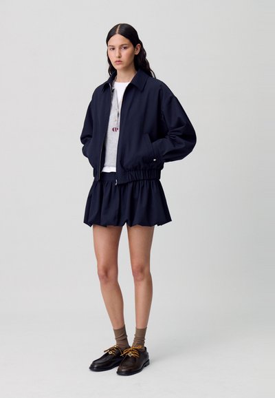 Blouson aviateur bleu marine avec ourlet élastique, associé à une jupe évasée assortie. Chemise grise unie en dessous et chaussures à lacets avec chaussettes marron.