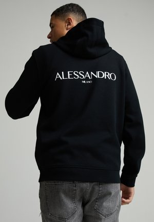 Junger Mann mit dem Rücken zur Kamera, der einen schwarzen Hoodie mit weiß gedrucktem "ALESSANDRO MILANO" auf dem Rücken und graue Jeans trägt.