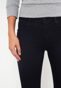 Jeans in denim blu scuro con vestibilità slim, piccole tasche frontali e chiusura a bottoni. Mano posata sulla vita, evidenziando la consistenza morbida.