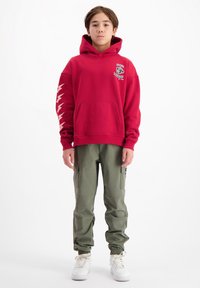 Rode hoodie met een voorzak en grafische print. Witte bliksempatronen op de mouwen. Olijfgroene cargobroek met elastieken manchetten. Witte sneakers.