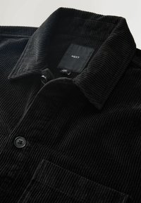 Schwarze Cordjacke mit umgeschlagenem Kragen, schwarzen Knöpfen und einem sichtbaren "NEXT"-Markenlabel im Nackenbereich.