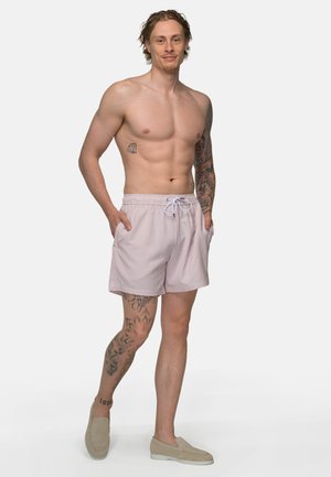 Man zonder hemd met tatoeages op linkerarm en rechterbeen, draagt lichtroze shorts met trekkoord en beige instapschoenen, staand met handen in de zakken.