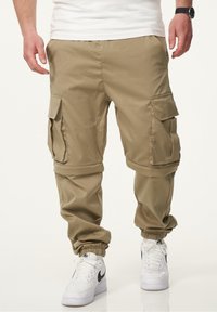 Khakifarbene Cargo-Hosen aus glattem Stoff, mit mehreren Seitentaschen, umgeschlagenen Knöcheln und einem lockeren Schnitt, kombiniert mit weißen Sneakers.