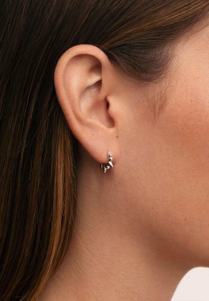 Gros plan sur l'oreille d'une femme aux cheveux bruns lisses, portant une petite boucle d'oreille argentée en forme d'anneau avec des accents en forme de pointe.