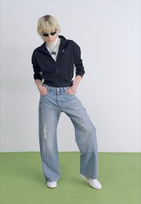Svart zip-up jacka, vit skjorta, ljusblå vida jeans med slitningar, vita sneakers och grön bakgrund med solglasögon som accessoar.