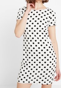 Bílé šaty s krátkým rukávem, které mají přiléhavý střih, černé puntíky a kulatý výstřih. Vyrobené z hladkého, pružného materiálu.