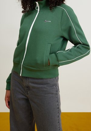 Veste zippée verte avec col montant, accents en piping blanc et logo Nike sur la poitrine. Comprend des poches latérales et des poignets côtelés.
