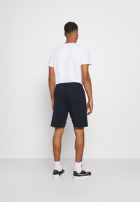 Marineblaue Shorts mit einem elastischen Bund und einer Gesäßtasche, kombiniert mit einem weißen T-Shirt mit kurzen Ärmeln und schwarzen Turnschuhen.
