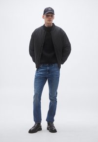 Μαύρο μπουφάν μπόμπερ πάνω από ένα σκούρο πλεκτό πουλόβερ, μπλε τζιν και μαύρα παπούτσια. Ένα μαύρο καπέλο με λευκό κείμενο ολοκληρώνει την εμφάνιση.