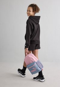 Mochila de tela rosa y azul, con un bolsillo delantero y un panel lateral de malla. Combinada con una sudadera negra y pantalones cortos negros.