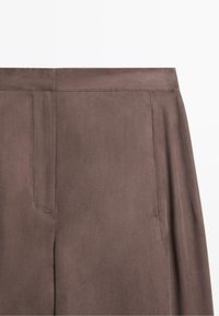 Nahaufnahme des Bunds und des oberen Oberschenkelbereichs einer braunen Hose mit Seitennaht und Taschen-Detail aus weichem Stoff.