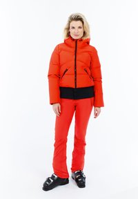 Mujer vestida con una chaqueta acolchada roja y pantalones, con botas de esquí negras, de pie contra un fondo blanco, mirando al frente.