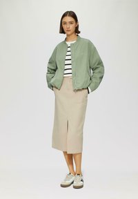 Veste bomber vert clair, chemise rayée blanche et noire, jupe midi beige avec fente avant, et baskets rétro blanches et brunes.