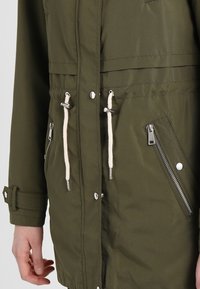 Oliivinvihreä takki, jossa on etupuolella napit, valkoiset nyörit, kulmikkaat vetoketjulliset taskut ja säädettävät hihansuut henkilön vartalolla.