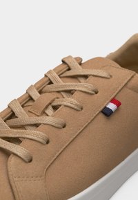 Κοντινό πλάνο σε μπεζ σουεντ αθλητικό παπούτσι με μπεζ κορδόνια και μια μικρή κόκκινη, λευκή και μπλε υφασμάτινη ετικέτα στην πλευρά κοντά στα κορδόνια.