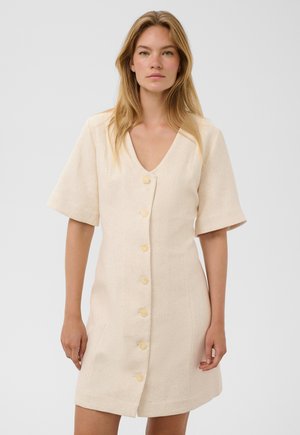 Femme aux longs cheveux blonds portant une robe beige à manches courtes et boutonnée, debout devant un fond clair uni.
