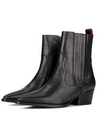 Botas de cuero negras hasta el tobillo con punta afilada, detalle de costura vertical y tacón bajo. Cuentan con una cremallera lateral y un tirador rojo.