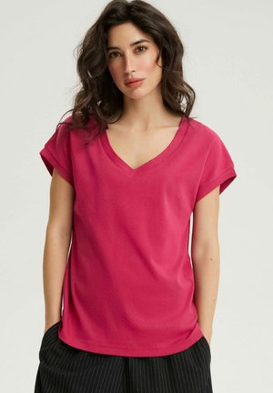 Kobieta o falujących ciemnych włosach, ubrana w jasnoróżowy top na krótki rękaw z dekoltem w serek oraz czarne spodnie w prążki, stojąca z rękami w kieszeniach.