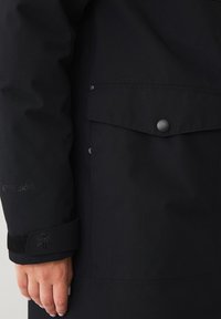 Giacca nera impermeabile con finitura testurizzata, dotata di tasca a flap con abbottonatura, chiusure a scatto e dettagli sui polsini in Velcro.