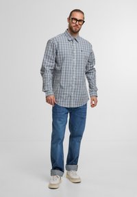 Mann mit Brille, der ein langärmliges, weiß-blau karriertes Hemd, eine blaue Jeans mit hochgekrempelten Säumen und weiße Sneakers vor einem schlichten Hintergrund trägt.