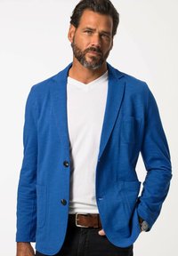 Blazer blu con una vestibilità rilassata, realizzato in tessuto testurizzato, con due bottoni frontali, una tasca sul petto e una maglietta bianca casual sotto.
