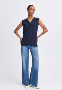 Débardeur marine sans manches avec un décolleté en V, détails de plis et trois boutons. Associé à un jean bleu à jambes larges et des escarpins blancs à bout pointu.