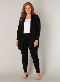 Zwarte lange cardigan over een witte top, gecombineerd met zwarte leggings en beige sandalen met bandjes. Gladde stof, open voorkant en slim fit.